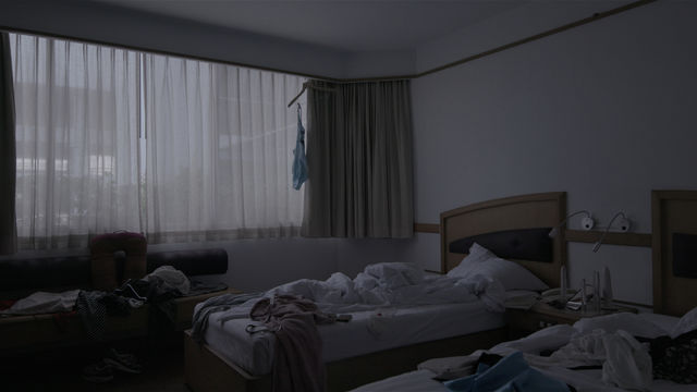 Image of a dark bedroom with two single beds that have messy white sheets. A large window in the background with sheet whitr curtains provides the only light.