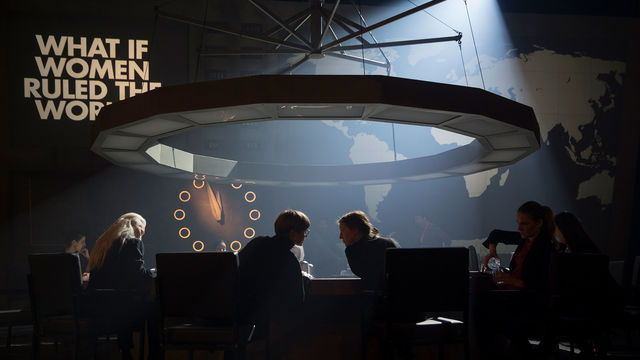 A circle of women around a table with a large circular light fixture above, and the words What if Women Ruled the World on the wall