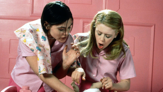 Two teenage girls in pink lean over a crib, holding a baby doll.