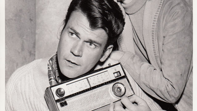 A man sits holding a radio, while a woman leans on him with a shocked expression.