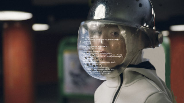 A woman wears a helmet with a plastic face shield. Formulaic text covers the face shield.