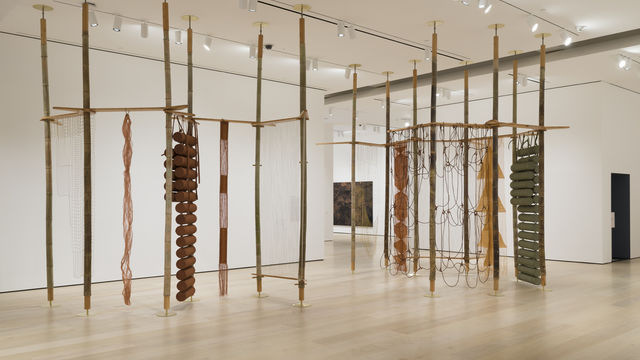 Gallery space with several long, wooden poles that reach from the floors to the ceilings adorned with woven strings and cylinders. 