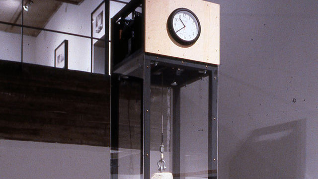 A large rock hung by a wire inside a clear glass box with a clock