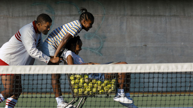 A man and a woman push a girl who sits inside of a shopping cart filled with tennis balls. 