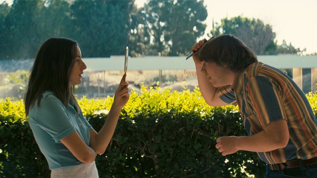 Girl and boy stand facing each other in front of a sunny hedge.