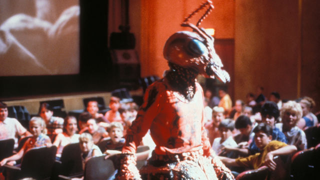 Person wearing a large bug costume walks around a crowded movie theater.