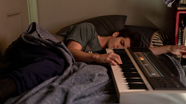 A man lies down on a bed with his fingers on a piano keyboard.