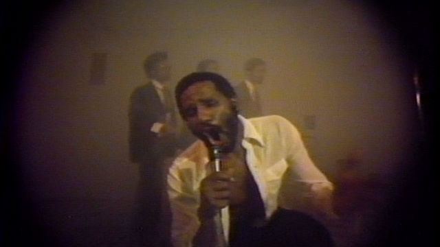 Grainy video still of artist Ulysses Jenkins singing into a microphone