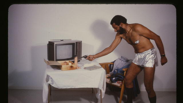 Photo of a man in silver shorts pointing a revolver at a dildo