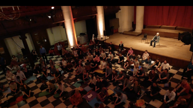 A person seated on an auditorium stage looking out over a small crowd seated on the floor