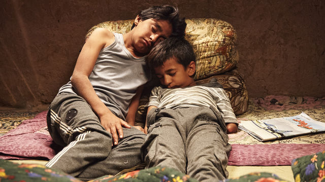 Two young boys lie together on several textiles and pillows.