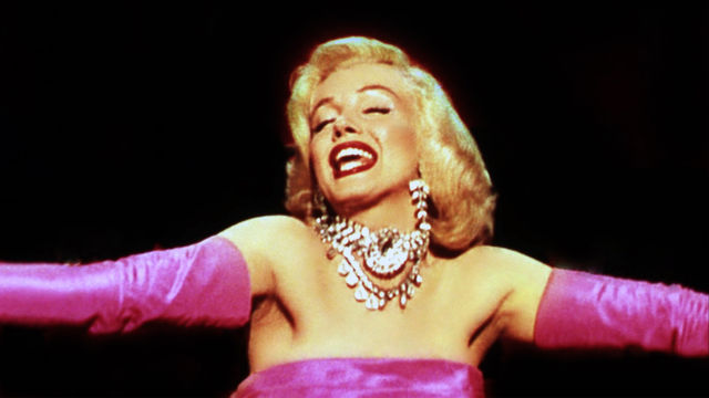 Marilyn Monroe with her arms spread out wearing a silky pink dress.