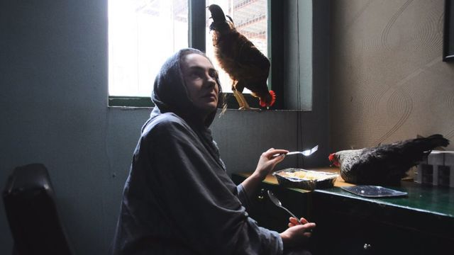 A woman sits at a desk with two chickens, eating her food. 