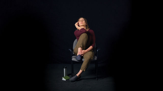 A woman sitting in an office chair holding her right knee up to her chest and her right hand to her face