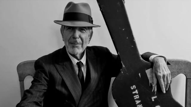 Black and white photo of musician Leonard Cohen seated with his arm around a guitar case