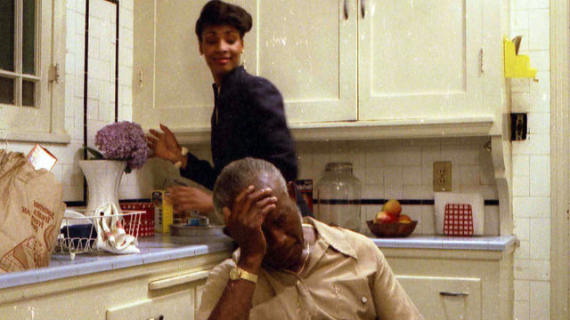 Man sits at a kitchen table, slumped over, while a woman is behind him.