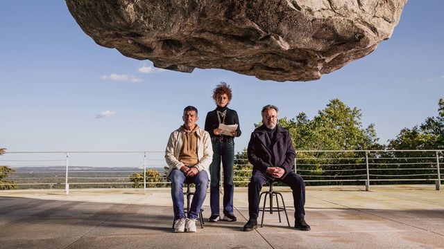 Film still from "Official Competition" (2022), showing a gigantic rock suspended over three people.