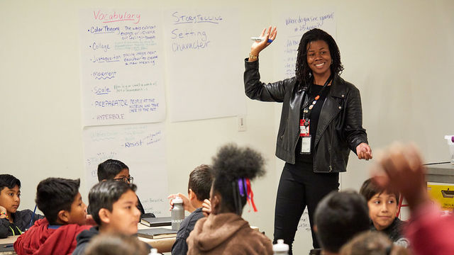 A woman with brown skin wearing all black holds her right arm up in front of a group of young children seated at tables covered in kraft paper.