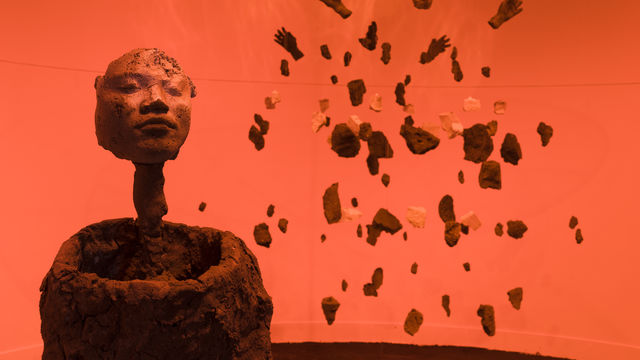 Photo of a sculpture of a form with a human face, and behind it an exploded view of a human figure suspended from the ceiling, in an orange-hued room