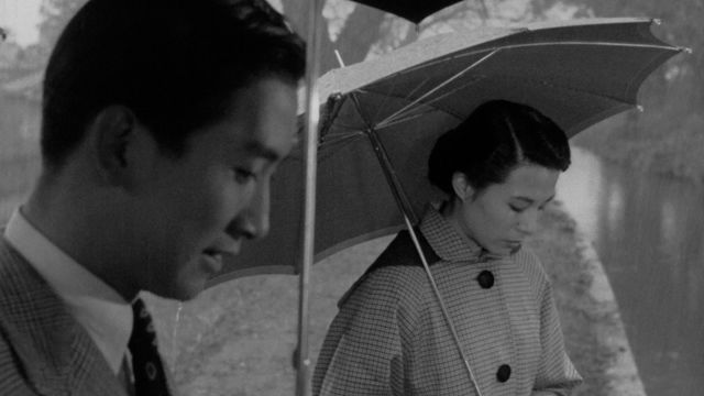 A man and woman hold umbrellas while looking at the river