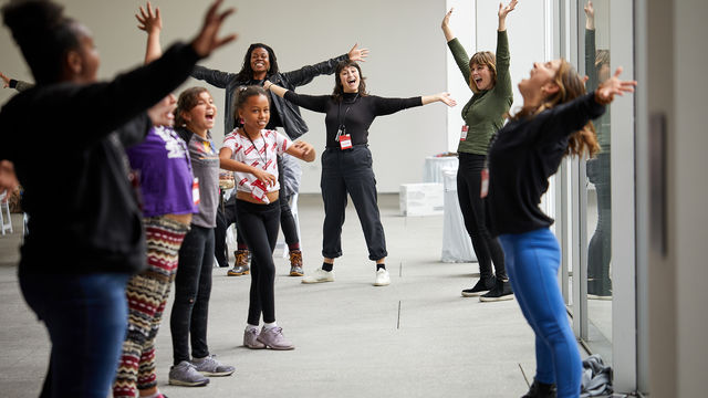 Seven people, young students on the left and educators in the center and on the right, stand with their legs wide and arms spread open smiling together. 