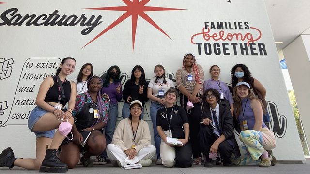 A group of young people stand, kneel, and site in front of a wall bearing painted signs of activist slogans ("ABOLISH ICE," Families belong together")