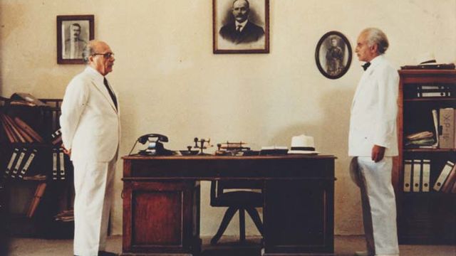 two men in white suits stare at each other across the length of a wooden desk