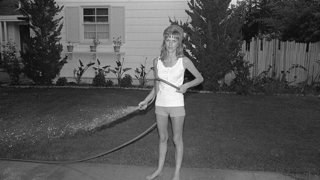 Black and white photo of a woman spraying the sidewalk with a hose