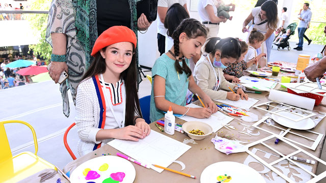 A girl with long brown hair wearing a red cap sits in front of a table with paint and miscellaneous craft items.