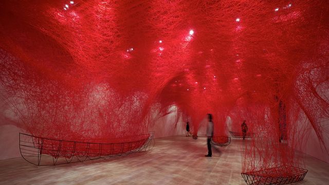 A person walking under an installation of fine red thread suspended from the ceiling and connect to small boat frames