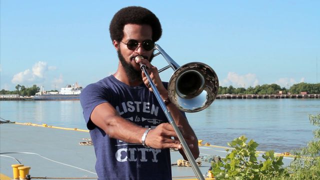 A Black man with an afro plays a trombone in front of a river