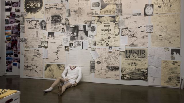 A gallery wall covered in drawings, with a headless mannequin sitting against it