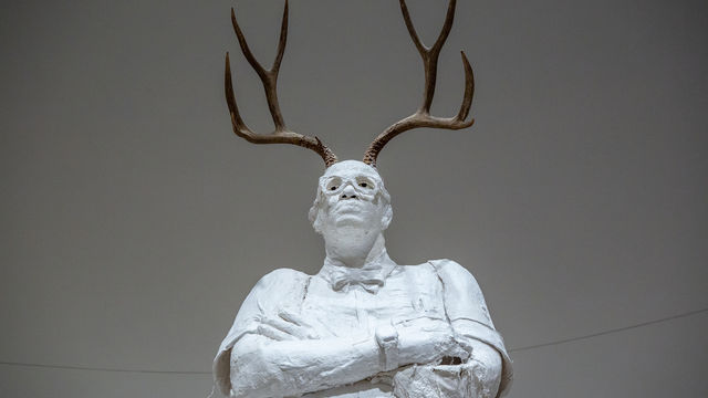 Life-size sculpture of a school principal standing with arms crossed, antlers growing from his head