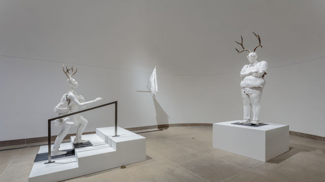 Life-sized white plaster figures of a girl climbing the stairs and a principal with arms crossed, both with antlers on their heads 