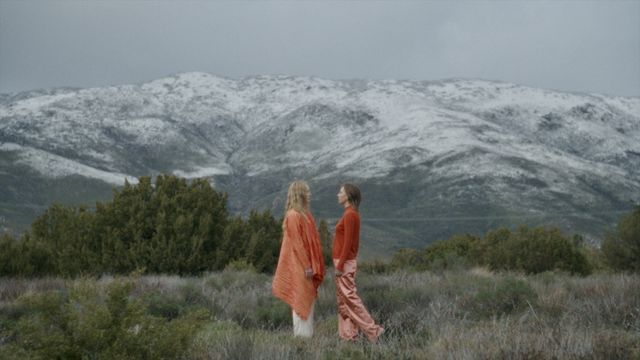 Two brightly dressed women stand facing each other in a field with a snow-capped mountain behind them