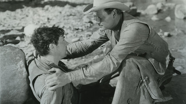 man with cowboy hat holds the shoulders of another man leaning against rock