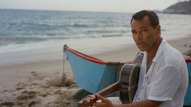 Rogê sitting with a guitar on a beach