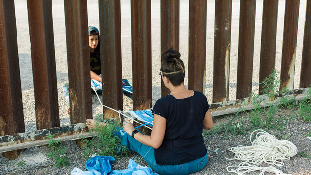 Two women sit on either side of the US-Mexico border fence, connected by a woven rope between them
