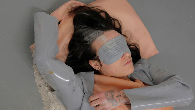 A man lays on a pillow, wearing a grey eye mask with text on it