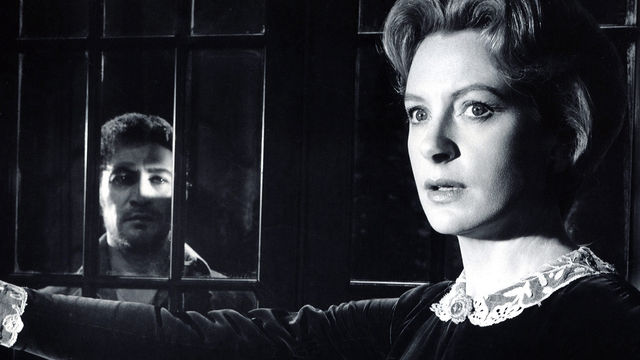A woman stands in the foreground and a man in the background, separated by prison bars