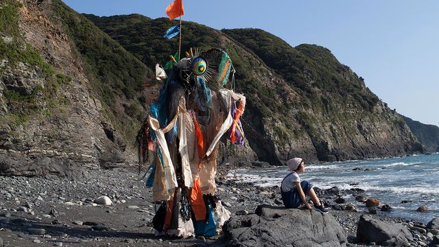 The artist Yangkura wearing a costume made of trash, standing on a beach