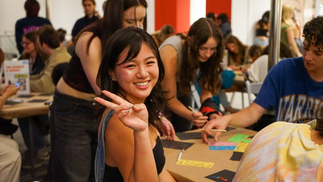 A young woman at an art making table smiles and makes a peace symbol with her finger