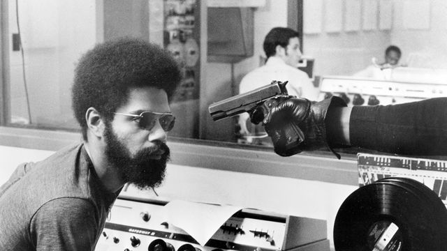 A gloved hand points a handgun at a Black man with a beard and afro