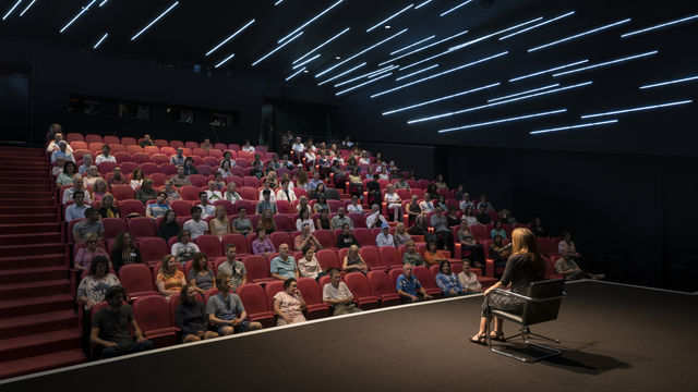An audience sitting in a darkened theater, watching a woman on stage