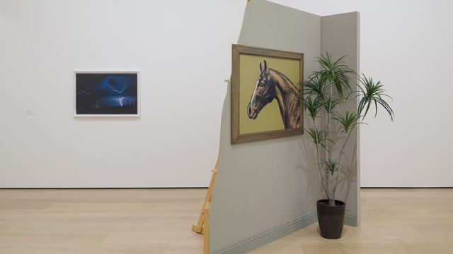 installation image with framed work on wall and a sculpture comprised of a false wall with painting and potted plant