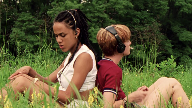 Two girls sit back-to-back in a field, wearing headphones