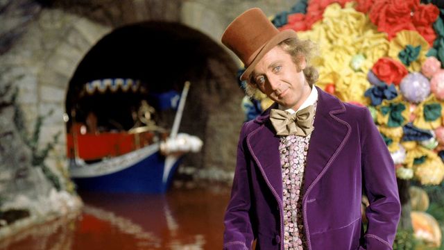 Willy Wonka wearing a brown top hat and purple coat stands in front of his chocolate river