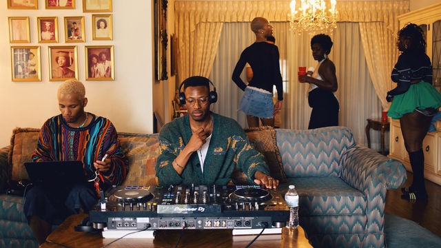 Two men sit at a couch behind a set of turntables, as partygoers gather around them