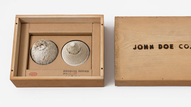 A simple wooden box: on the left, two small mounds of sand sit in the box, with a stamp reading "EROSION SERIES," on the right, the box top with the words "JOHN DOE CO." burned into it