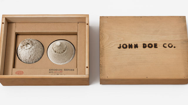 A simple wooden box: on the left, two small mounds of sand sit in the box, with a stamp reading "EROSION SERIES," on the right, the box top with the words "JOHN DOE CO." burned into it